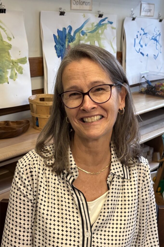 A figure sits in a warmly lit room, smiling, with long hair partially tucked behind their ear. They wear a patterned black and white shirt with a loose collar, complemented by simple jewelry. In the background, colorful children's artwork is displayed on the walls, featuring abstract smudges of blue and green paint. Wooden tables and various items suggest an artistic environment, contributing to a vibrant and creative atmosphere.