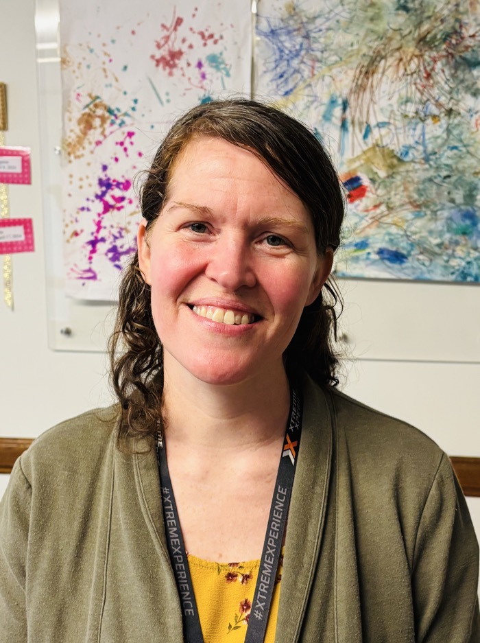 A person smiles directly at the viewer, framed by a soft, neutral wall. Their attire includes a muted olive-green cardigan over a bright yellow floral shirt. Behind them, colorful abstract artworks featuring splashes of pink, blue, and gold create a vibrant backdrop. The lighting is warm and even, enhancing the convivial atmosphere of the setting.