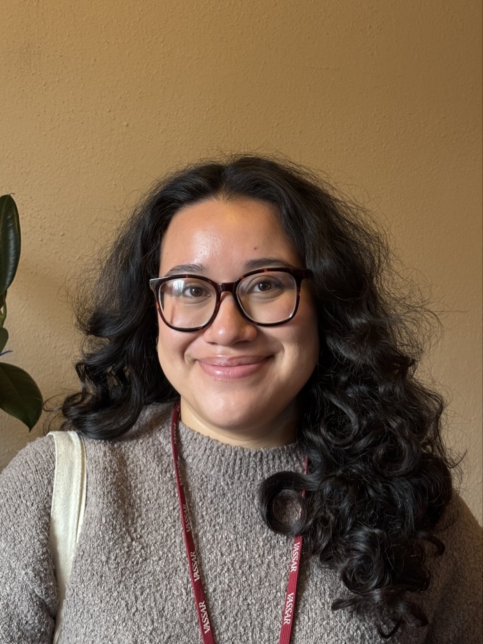 A person stands against a textured, warm-toned wall, illuminated by soft, natural light. They have curly, dark hair that falls loosely around their face and are wearing large glasses. A cozy, light gray sweater contrasts against the earthy backdrop. In the foreground, a green plant adds an organic element to the composition, enhancing the atmosphere of the setting.