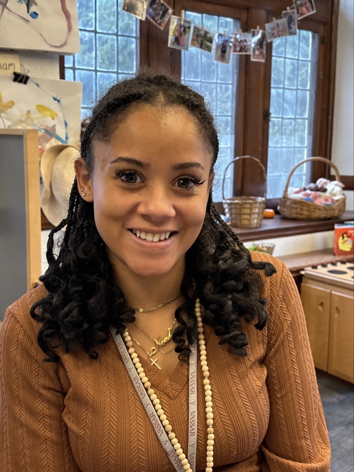A person smiles in a warmly lit indoor environment, featuring wooden cabinetry and soft, natural light filtering through large windows. The individual has curly hair adorned with jewelry and is wearing a ribbed, brown top, which contrasts with the light-colored backdrop. In the background, various colorful artworks and photographs are displayed, adding visual interest to the setting. The overall composition conveys a sense of creativity and warmth within a welcoming space.