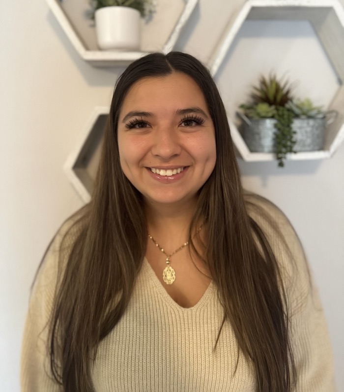 A person stands smiling in front of a light-colored wall adorned with hexagonal shelves. The individual wears a beige, textured sweater and a delicate necklace. Above, the shelves hold small potted plants, contributing to a natural aesthetic. The soft indoor lighting enhances the overall warmth of the scene, creating an inviting atmosphere.