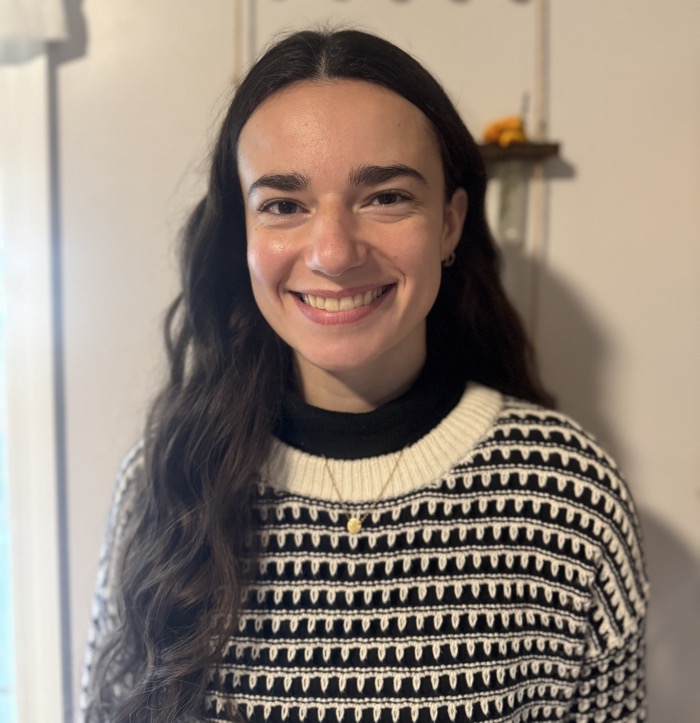 A person is smiling, facing the camera with a pleasant expression. They are wearing a black and white patterned sweater with a high collar and layered underneath a black top. The background features a light-colored wall with a small shelf displaying bright objects, contributing to a well-lit atmosphere. Soft, natural light enhances the warmth of the scene, creating a welcoming environment.