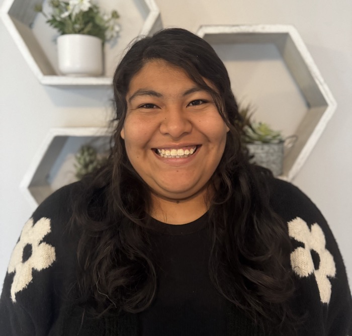 A smiling figure is centered within the image, wearing a black sweater adorned with large white floral patterns. The background features a light-colored wall with hexagonal shelves containing small plants and decorative items, creating a structured contrast. Soft, diffused lighting illuminates the scene, enhancing the warm, inviting atmosphere. The overall composition is balanced and symmetrical, drawing focus to the figure in a friendly and welcoming manner.