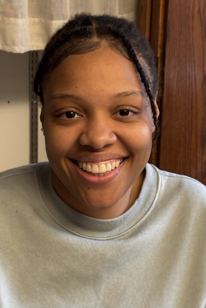 A person smiles directly at the camera with a broad smile. Their hair is styled in braids, with some sections pulled back, while they wear a light blue sweatshirt. The background features a wooden surface partially obscured by soft, off-white fabric. Soft indoor lighting creates a warm atmosphere, highlighting the facial features and the texture of the sweatshirt.