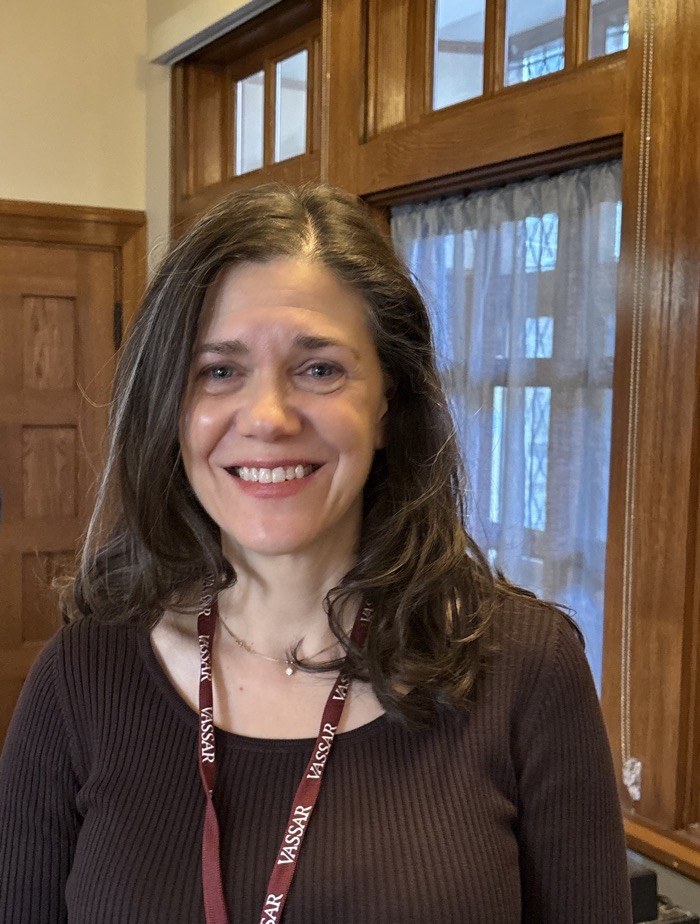 A person with long, dark hair stands in a wood-paneled room. They are smiling at the camera, and have a maroon lanyard around their neck with the word "Vassar" repeated on it in white lettering. They are wearing a brown shirt.