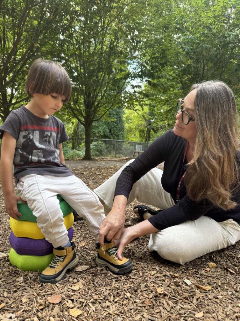 A figure sits on a vibrant, multi-colored stack of padded shapes, wearing a dark graphic t-shirt and light pants, with their head down, focused on their footwear. Another figure kneels beside them on a textured brown wood chip surface, adjusting the laces of sturdy yellow boots. Surrounding the scene is lush greenery with sunlight filtering through the leaves, creating dappled shadows on the ground. The atmosphere is casual and engaging, showcasing an interaction in a natural setting.
