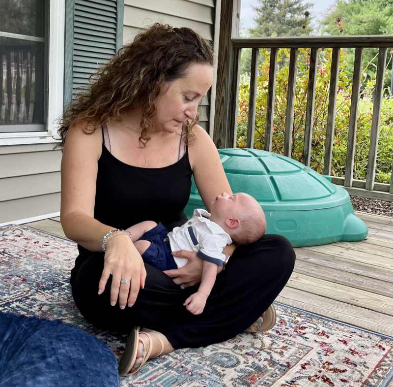 A figure sits on a patterned rug, wearing a black outfit, with one arm cradling an infant dressed in a white shirt and dark shorts. The figure leans slightly forward, gazing down at the infant, who is looking up with a relaxed expression. In the background, a green play structure is partially visible, surrounded by lush foliage that hints at an outdoor setting. The scene is softly lit, suggesting an overcast sky, with natural textures from the wooden porch and greenery enriching the composition.