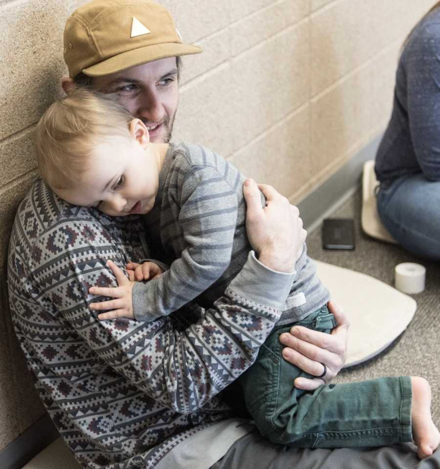 A figure sits on a floor covered in gray carpeting, holding a small child closely. The adult wears a patterned sweater and a cap with triangular embellishments, while the child leans against them, dressed in a striped shirt and dark pants. In the background, a textured wall provides a neutral backdrop, and a few other figures are visible, engaged in various activities. Soft lighting fills the space, creating a calm and inviting atmosphere.